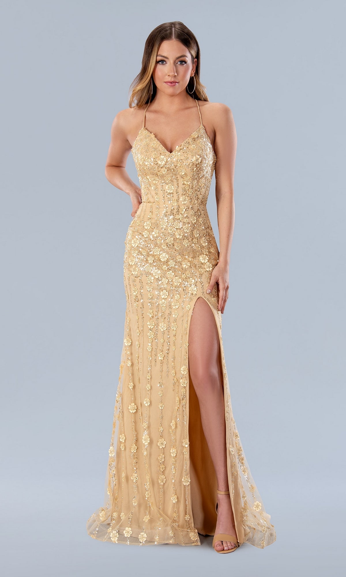 A woman models the Long Prom Dress 24189 by Stella Couture—a floor-length, gold, sleeveless fitted gown with sequin floral patterns, spaghetti straps, lace-up back, and a thigh-high slit—against a plain light gray background.